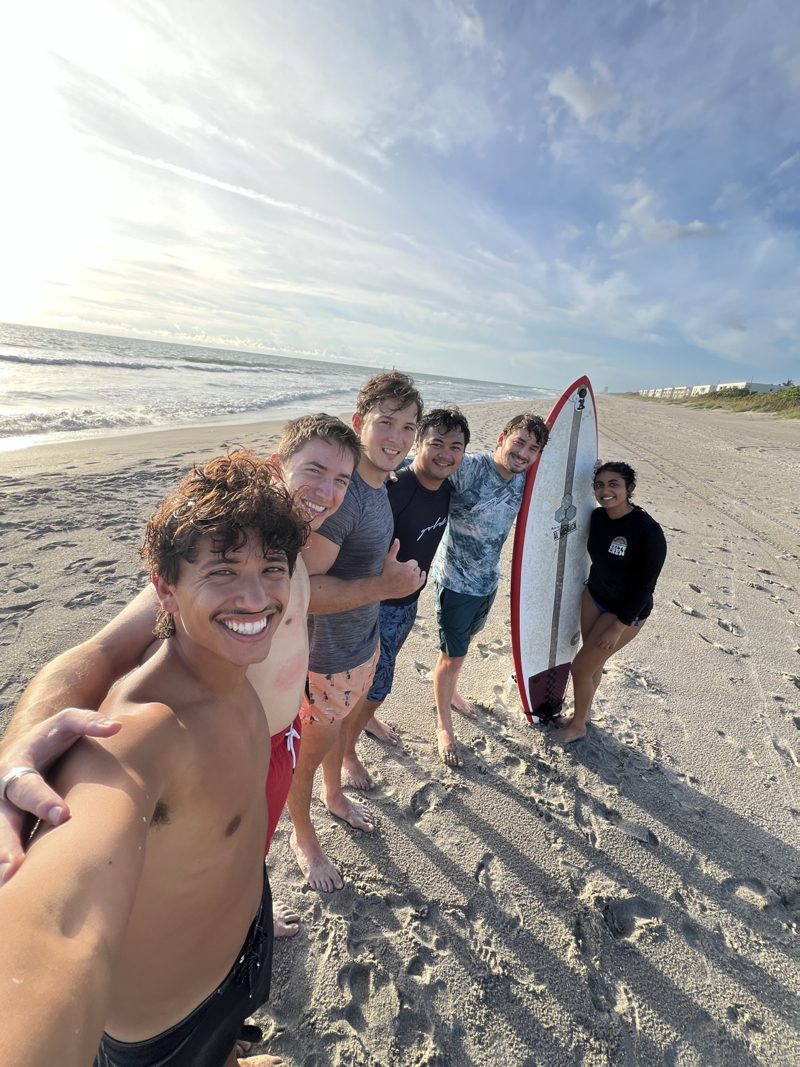 Beach Medicine Club members smiling together on the beach with a surfboard