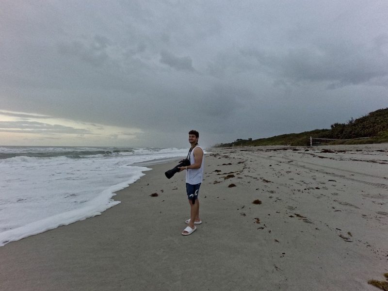 Tryfon carrying a camera along the shoreline beneath a cloudy sky
