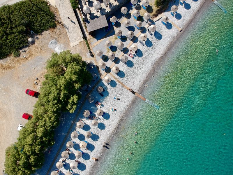 Aerial view of beach umbrellas and swimmers along a Greek shoreline with clear turquoise water